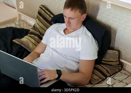 Casual young adult male working remotely on a laptop from comfortable bed setting indoors Stock Photo