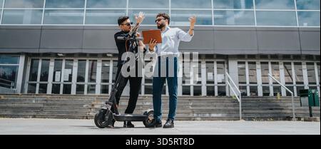 Two colleagues discussing work details while using an electric scooter outdoors. Stock Photo