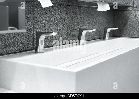 Row of three modern chrome faucets over a long white rectangular sink against a textured gray granite wall in a public restroom. High quality photo Stock Photo