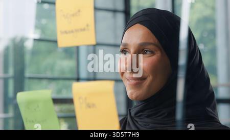 Satisfied islamic business woman analyzing stickers on glass in office Muslim businesswoman in hijab company CEO entrepreneur female girl reading Stock Photo