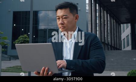 Asian middle-aged businessman typing laptop in city Korean business man serious browsing wireless computer in downtown internet virtual job work task Stock Photo