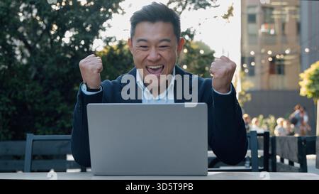Happy excited Asian business man winning with laptop rejoice looking computer Korean businessman win achieve victory online internet job working in Stock Photo