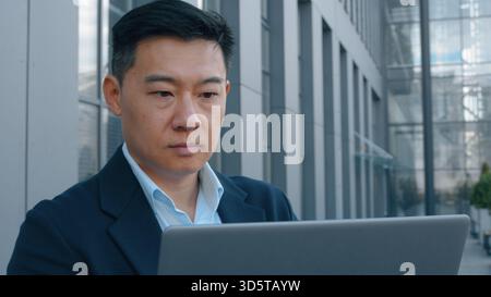 Serious Asian middle-aged business man with laptop in city working focused Korean businessman Chinese Japanese entrepreneur employer financial analyst Stock Photo