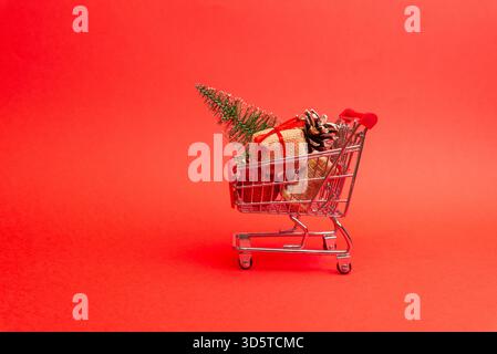Mini shopping cart with Christmas gifts, pine cones and decorations on a clean red background for holiday themes Stock Photo