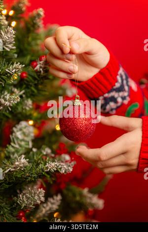 Closeup of hands placing a red Christmas ornament on a decorated tree with lights, berries, and snow-tipped branches Stock Photo
