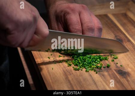 A person is cutting green herbs on a wooden cutting board. The knife is silver and the herbs are chopped into small pieces Stock Photo