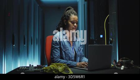 Sitting tech in denim shirt and lanyard at server aisle, using laptop and yellow Ethernet cable Stock Photo