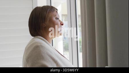 Standing mature woman in light knit cardigan, looking out large glass window at home with shutters Stock Photo