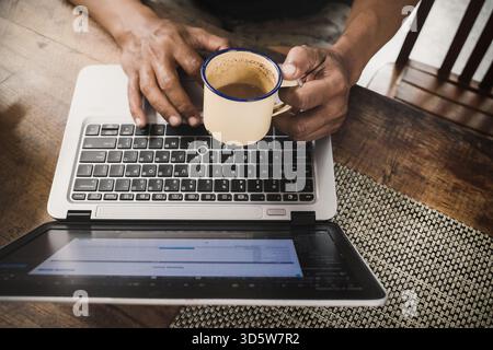 Asian adult male person working his freelance business on a notebook computer at home with one hand holding a cup of coffee. work from home concept. Stock Photo