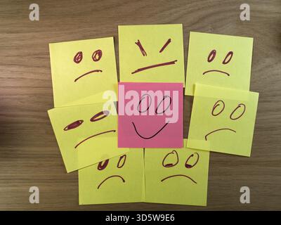Happy Face Among Sad, Angry Faces - Pink sticky note with happy face surrounded by yellow notes with sad, angry faces on wood. Emotions, mental health Stock Photo