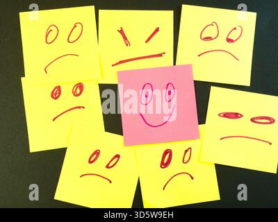 Happy Face Stands Out in Crowd of Sad - One pink sticky note with a happy face stands out from many yellow sticky notes displaying sad and angry expre Stock Photo