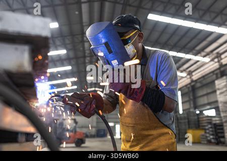 A worker engaged in welding inside a large industrial workshop. The individual is wearing protective gear including a welding helmet, gloves, and apro Stock Photo
