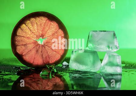 A citrus slice glows against green, paired with stacked ice cubes on a reflective surface. Stock Photo