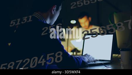 Working Asian man in dark sweater typing on laptop in dim office, with numeric overlays Stock Photo