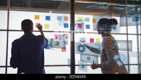 Writing women colleagues in jackets sketching on glass partition at office, with sticky notes, code Stock Photo