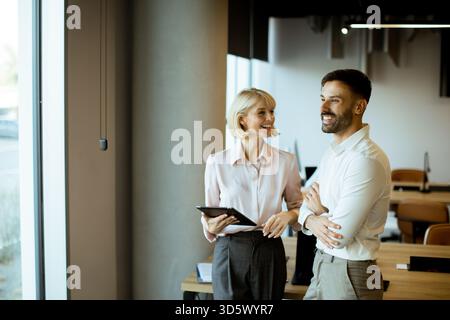 Two professionals engage in a lively discussion in a contemporary office. Natural light floods the space, highlighting their friendly expressions and Stock Photo