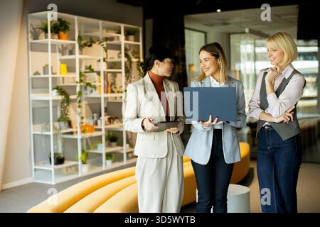 Three women engage in a lively conversation in a modern office. They hold a laptop and a tablet, discussing plans in a vibrant workspace filled with p Stock Photo