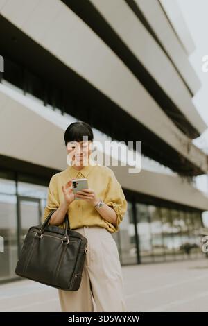 A woman stands outside a contemporary building, checking her cellphone with a smile. She wears a yellow shirt and beige pants, holding a black bag, en Stock Photo