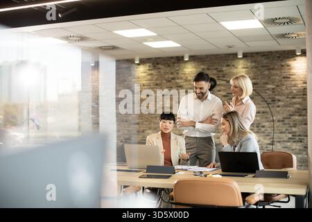Group of diverse professionals engaged in a meeting, discussing ideas by a table with laptops, in a well-lit office with stone walls. They appear focu Stock Photo