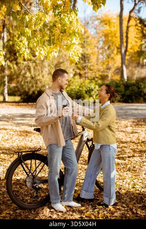 A Korean woman and a Caucasian man share a joyful moment in a vibrant autumn park, surrounded by falling leaves and their bicycles. Stock Photo