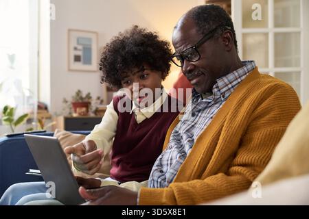 Senior Black Man and Teenager Boy Using Digital Tablet Together Indoors Stock Photo