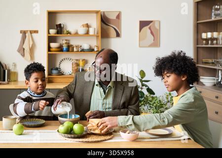Senior Black Man Having Breakfast with Two Black Boys in Kitchen Setting Stock Photo