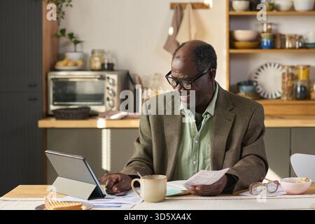 Senior Black Man Using Digital Tablet While Reviewing Financial Documents Stock Photo