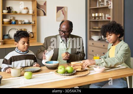 Senior Black Man Pouring Milk for Two Black Boys Having Breakfast Together Stock Photo