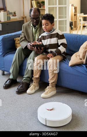 Senior Black Man Teaching Black Boy Using Tablet with Robot Vacuum Stock Photo