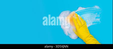 Hand in Yellow Glove Holds Sponge and Wipes Soapy Foam on Blue Background Stock Photo