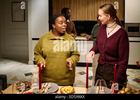 Young adult Black woman and young adult Caucasian woman standing at dining table talking, multiethnic male friends in background preparing food during Thanksgiving dinner among neighbours Stock Photo