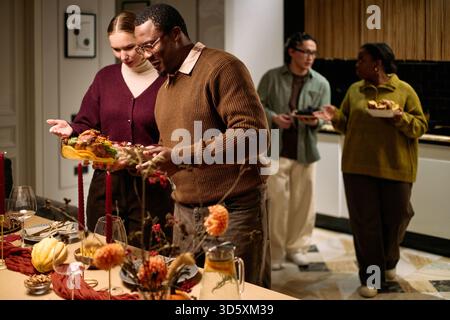 Multiethnic group of young adults and middle aged people serving traditional Thanksgiving dinner, Caucasian woman and Black man placing food on table while Asian man and Black woman talking Stock Photo