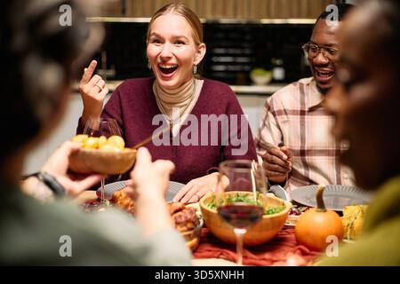 Diverse group of young adults and middle aged people laughing and sharing food around table during Thanksgiving dinner, Caucasian woman smiling, Black man enjoying meal, hands passing dishes Stock Photo