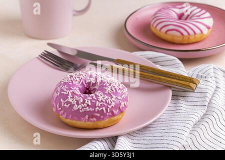 Two sugar-coated donuts lie on a pink ceramic plates. Serving with cutlery, mug and linen napkin. Selective focus Stock Photo