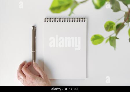 Top view mockup of an open blank notebook on spiral, leaves of houseplant and an automatic pen adjusted by female hand. Selective focus, soft focus. C Stock Photo