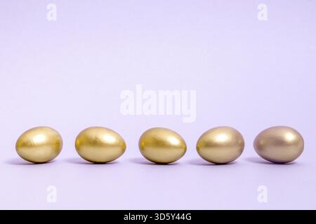 Five Easter eggs decorated with gold paint lie in a row against a lavender background. Concept for Easter, spring. Selective focus. Copy space Stock Photo