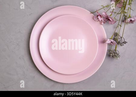 Close-up flat lay of empty pink plate and sprig of pink wildflowers on rough gray concrete or cement background. Spring concept. Selective focus Stock Photo