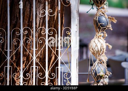 A close-up image showing a white, ornate, and rusty metal gate next to a tree trunk, with decorative black and white stones hanging in a woven net. Stock Photo