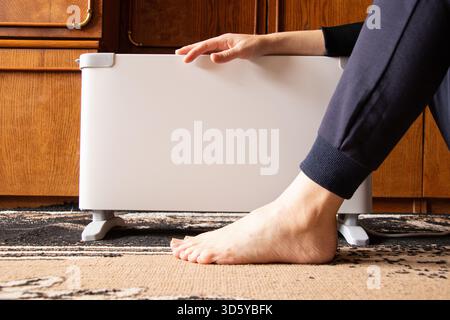 A girl sits near an electric convector on a carpeted floor in a residential apartment, warming herself, against a backdrop of wooden furniture in Ukra Stock Photo