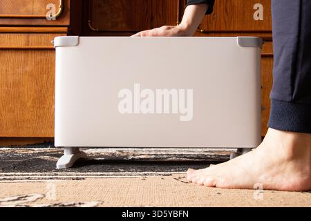 A girl sits near an electric convector on a carpeted floor in a residential apartment, warming herself, against a backdrop of wooden furniture in Ukra Stock Photo