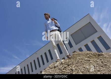 Business man standing on pile of gravel, heap of gravel, in front of office building, construction site manager, supervisor, supervision, worker, work Stock Photo