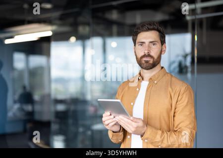 Young businessman holding a digital tablet with a serious expression, looking directly at the viewer, standing in a contemporary office representing digital communication and business technology Stock Photo