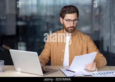 Businessman reading paper documents and reviewing reports while working on a laptop at a contemporary office desk, demonstrating dedication and success in his career Stock Photo