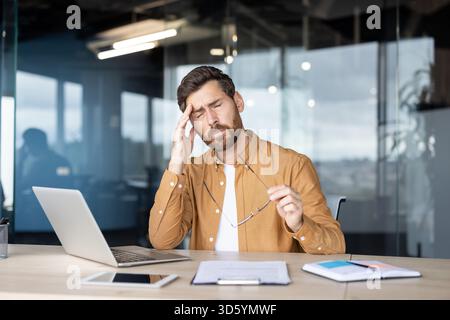 Businessman feeling severe headache while touching his temple, experiencing stress, tiredness, and burnout from excessive work at a contemporary glass office Stock Photo