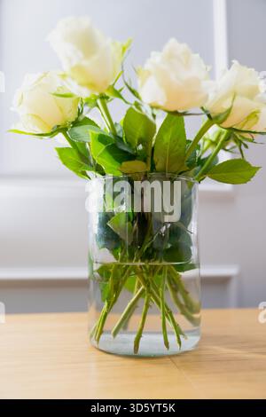 Fresh white rose bouquet placed in transparent glass vase showing layered leaves, submerged stems and soft natural tones ideal for home decoration Stock Photo