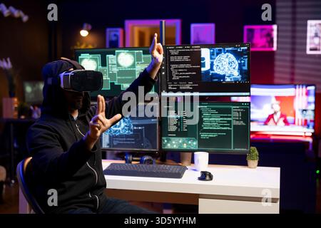 Hacker installing spyware using virtual reality tech and AI to secretly gather data about user activity. Rogue developer coding exploits using VR headset to gain unauthorized access Stock Photo