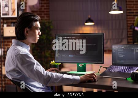 Self employed man sitting indoors during evening hours, concentrating on multiple screens, working on html codes. Male asian programmer reviewing online applications to develop new website ideas. Stock Photo