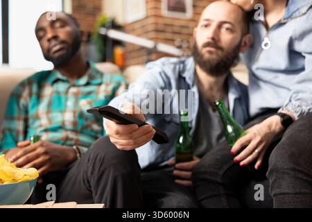 Close up of caucasian man using remote to switch channels on television. White bearded man adjusts volume and changes shows while friends sit nearby on couch during casual home hangout. Stock Photo