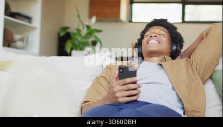 Lying man wearing tan shirt and blue pants, holding smartphone, black headphones on light sofa Stock Photo