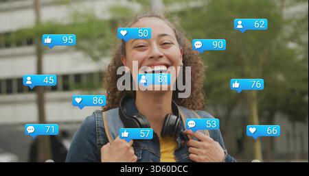 Smiling woman in denim jacket on campus holding backpack with over-ear headphones amid blue bubbles Stock Photo